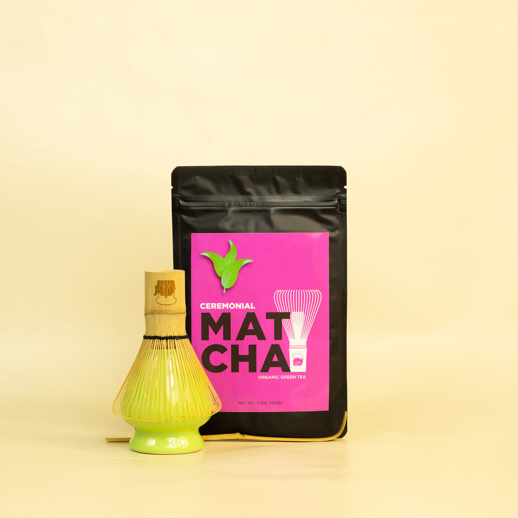 A bag of ceremonial matcha, a whisk, a whisk stand, and a matcha spoon against a yellow background