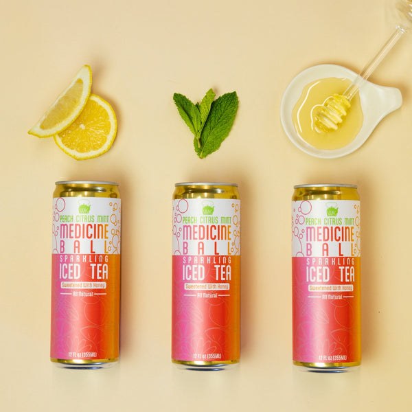 Three cans of Medicine Ball Iced Tea on a beige background with lemon slices and a spoonful of honey.