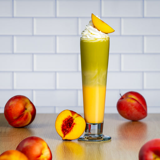 Peach matcha latte layered with creamy milk, topped with whipped cream and a fresh peach slice, surrounded by ripe peaches