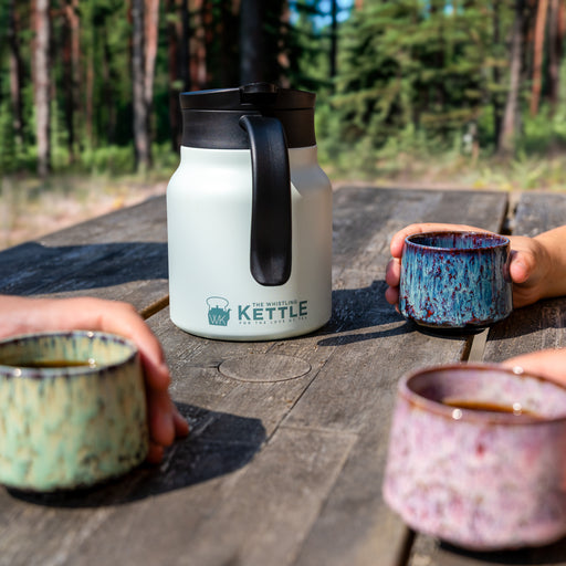 Three people enjoy tea around a picnic table, with a teal insulated tea carafe and colorful ceramic cups in a woodsy outdoor setting