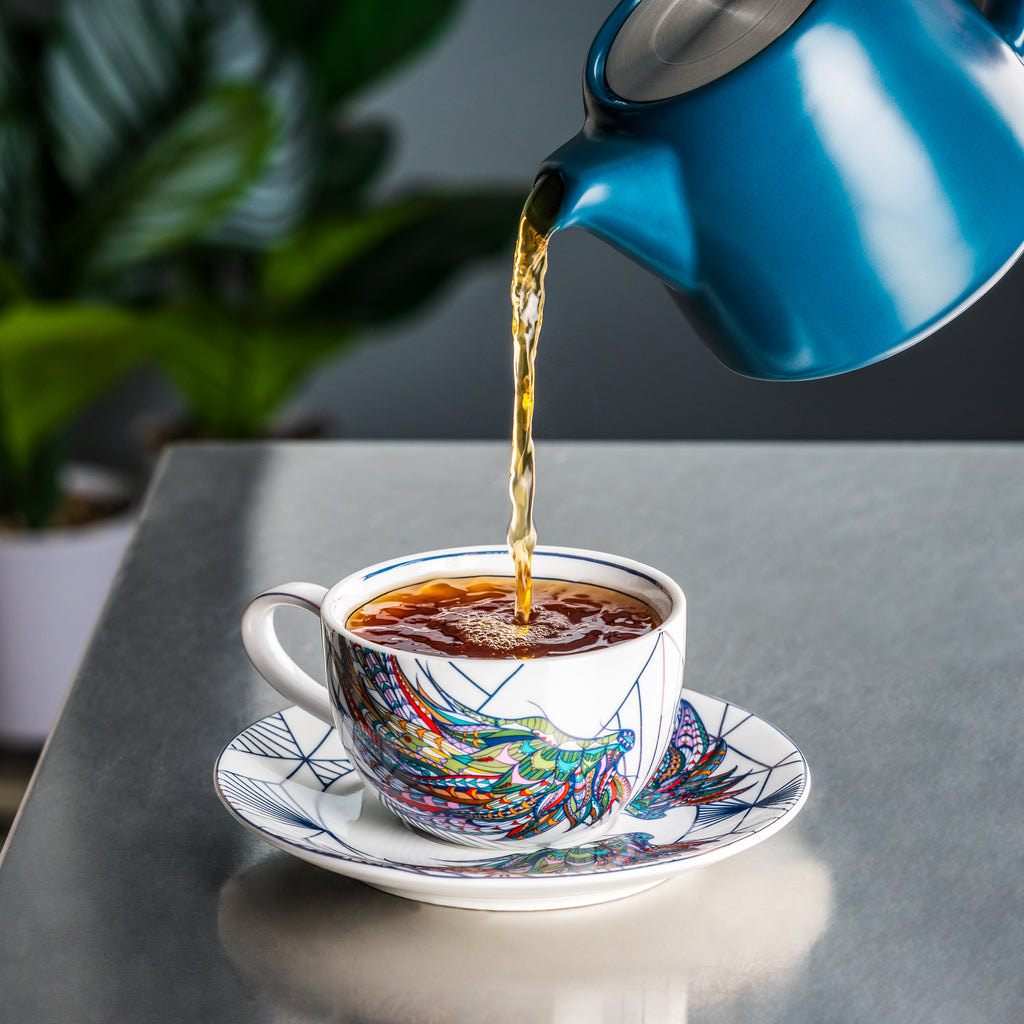Blue teapot pouring tea into a colorful teacup on a gray surface with a plant in the background.