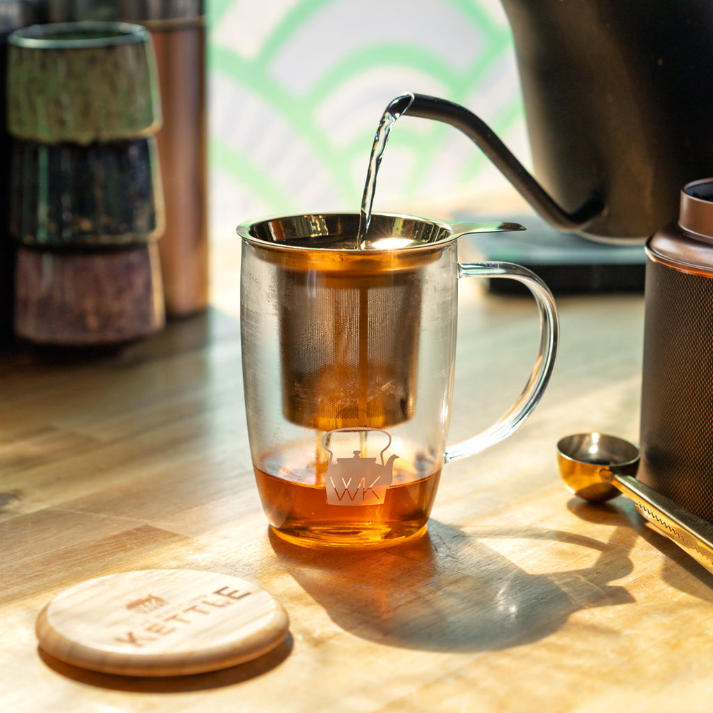 Hot water pours from a gooseneck kettle into a glass tea mug with a stainless steel infuser, surrounded by tea accessories in warm sunlight