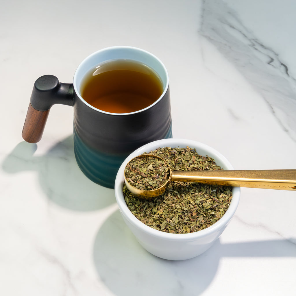 Freshly brewed Spearmint Tea in a Wave Mug beside loose-leaf spearmint tea and a golden scoop on a bright marble surface.
