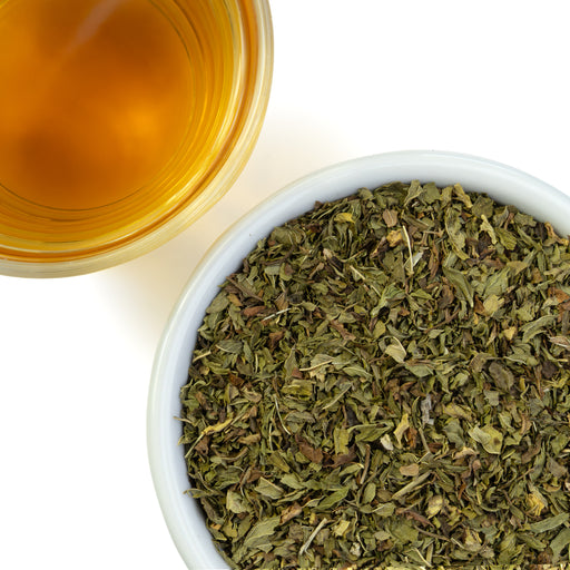 Spearmint leaves in a bowl next to a glass of brewed tea on a white background.