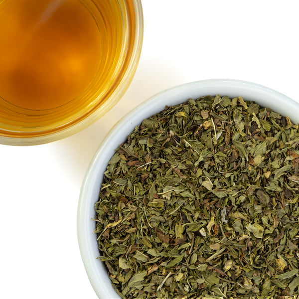 Spearmint leaves in a bowl next to a glass of brewed tea on a white background.