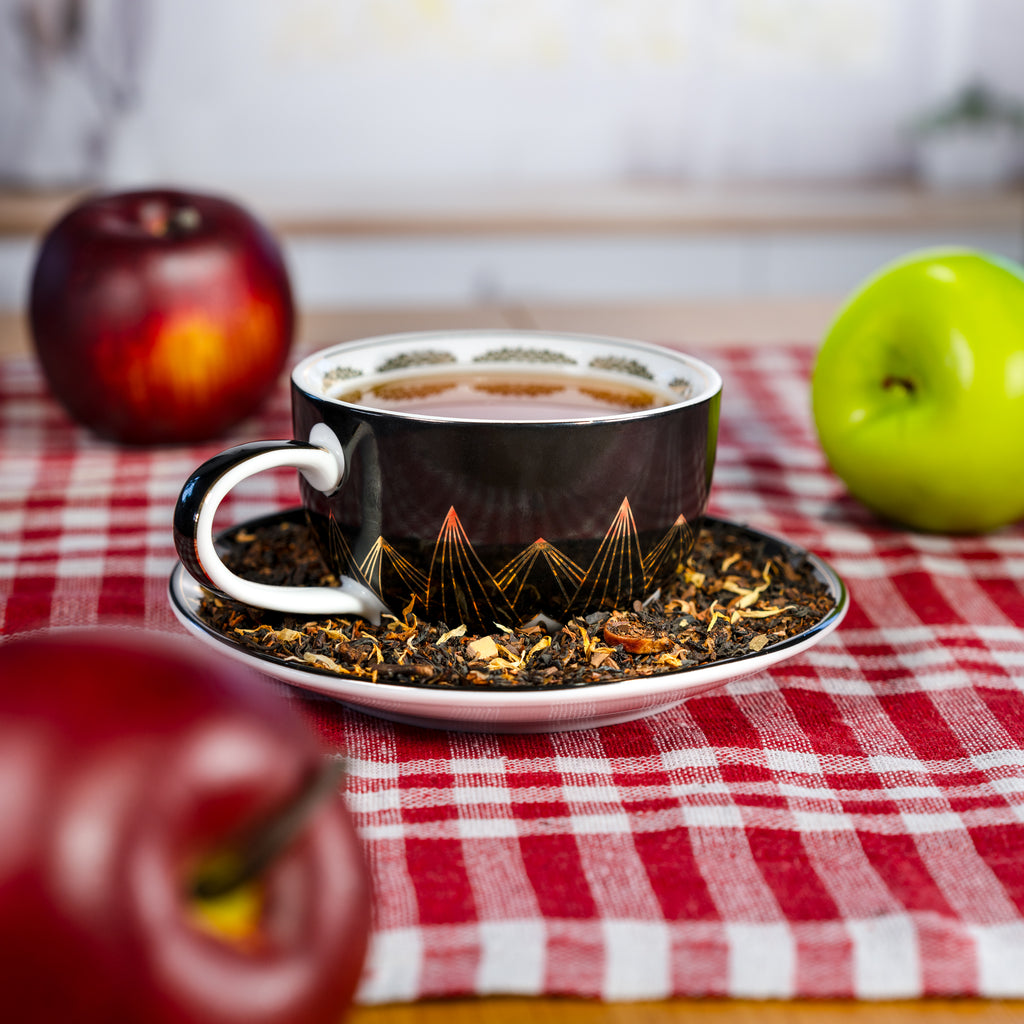 Teacup with saucer on a red and white checkered tablecloth with apples around.