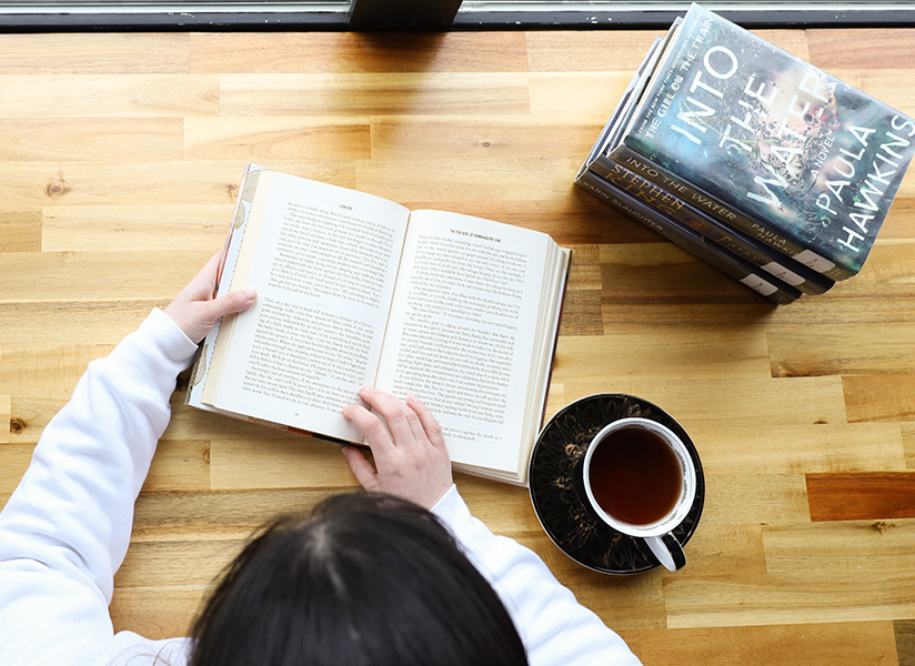 Top-down view of a woman reading a book with a cup of tea at her side.