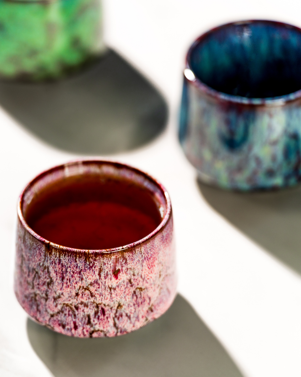 Three ceramic cups with unique glaze patterns on a reflective surface.