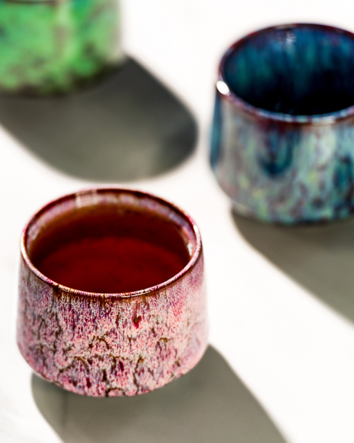 Three ceramic cups with unique glaze patterns on a reflective surface.