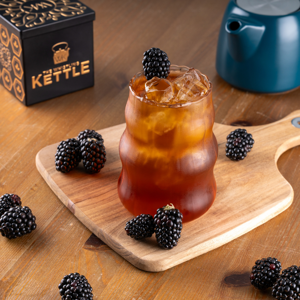 Iced tea with blackberries on a wooden cutting board, The Whistling Kettle brand is visible on a stylized tea tin.