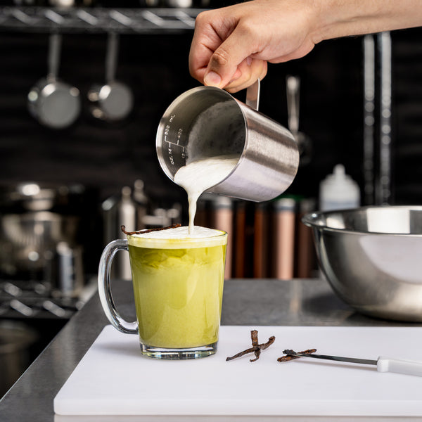 Vanilla matcha latte topped with steamed milk being poured into a glass mug, garnished with a vanilla bean