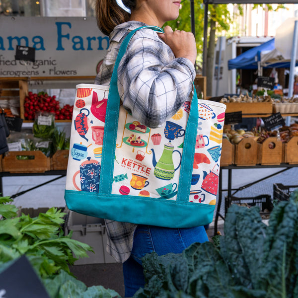 A woman at an outdoor farmers market carries a white canvas tote bag with a colorful tea-themed print and teal trim. The bag features illustrations of teapots, cups, mugs, and a kettle, with the word 'KETTLE' printed on the front.