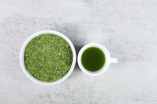 A cup of brewed Yuzu Matcha Kukicha next to a bowl of loose leaf tea leaves